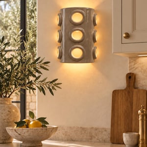 Op de afbeelding: Een halfcilindervormige wandlamp met een glanzende taupekleurige afwerking, voorzien van drie verticale ronde uitsparingen aan de voorzijde en twee aan elke zijkant, die een warme, zachte gloed van binnenuit verspreidt.