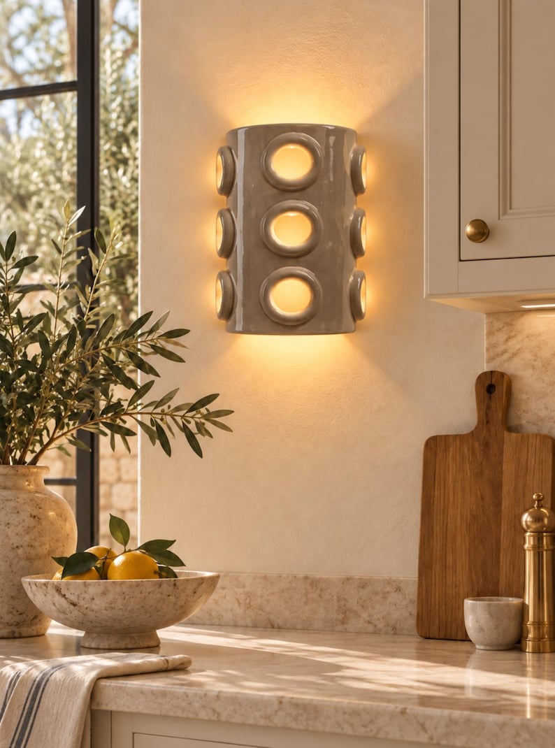 Op de afbeelding: Een halfcilindervormige wandlamp met een glanzende taupekleurige afwerking, voorzien van drie verticale ronde uitsparingen aan de voorzijde en twee aan elke zijkant, die een warme, zachte gloed van binnenuit verspreidt.