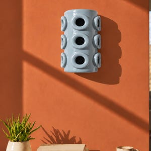 Op de afbeelding: Een cilindervormige wandlamp van lichtblauw keramiek met drie verticale rijen ronde openingen. Het armatuur heeft een gladde, glanzende afwerking en een holle binnenkant, ontworpen om vlak tegen een verticaal oppervlak te worden gemonteerd.