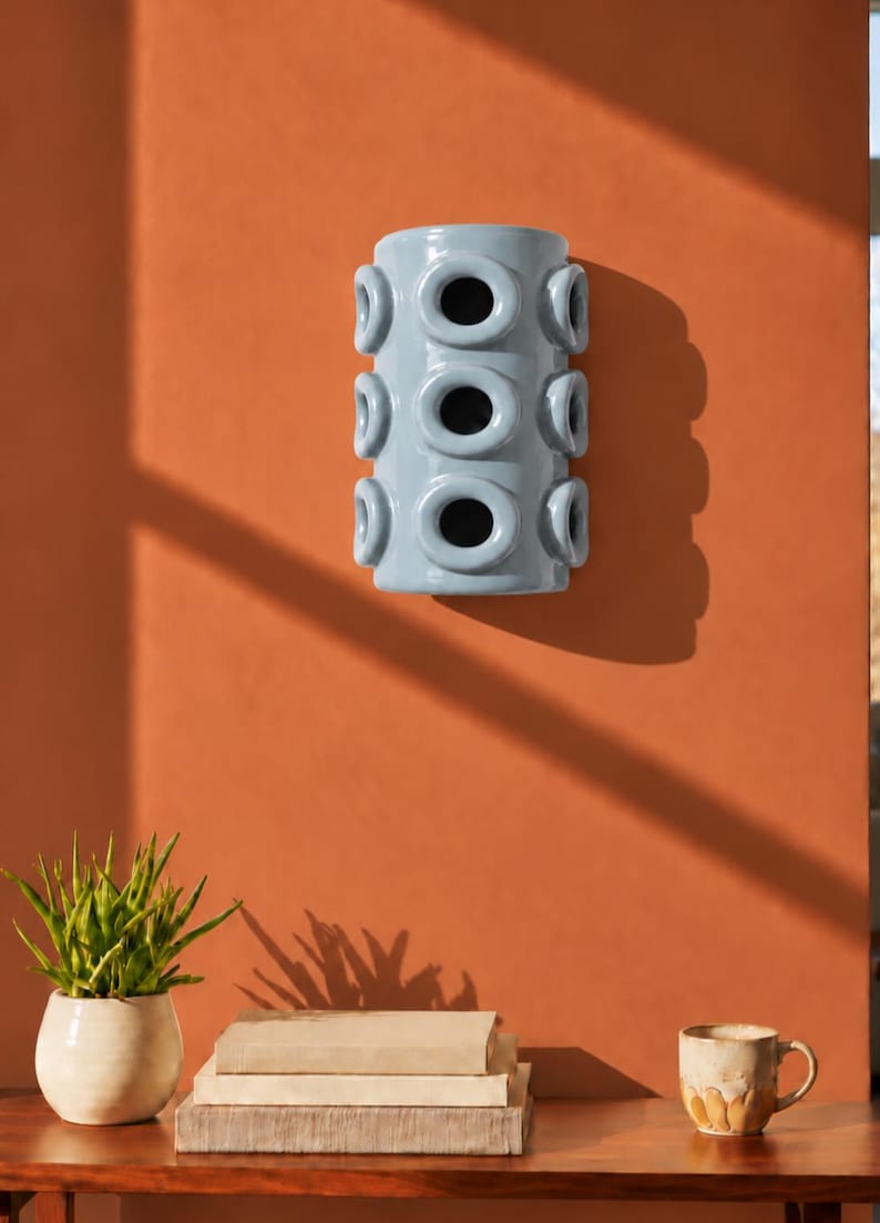 Op de afbeelding: Een cilindervormige wandlamp van lichtblauw keramiek met drie verticale rijen ronde openingen. Het armatuur heeft een gladde, glanzende afwerking en een holle binnenkant, ontworpen om vlak tegen een verticaal oppervlak te worden gemonteerd.