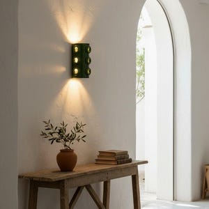 Op de afbeelding: Een donkergroene wandlamp met zes ronde glazen lenzen gemonteerd op een witte muur, die warm licht naar boven en beneden uitstraalt. Het armatuur heeft een verticaal rechthoekig lichaam met drie paren cirkelvormige openingen.
