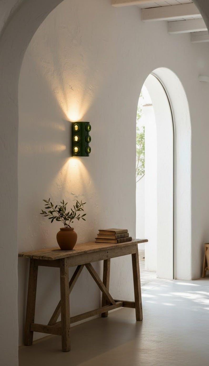 Op de afbeelding: Een donkergroene wandlamp met zes ronde glazen lenzen gemonteerd op een witte muur, die warm licht naar boven en beneden uitstraalt. Het armatuur heeft een verticaal rechthoekig lichaam met drie paren cirkelvormige openingen.