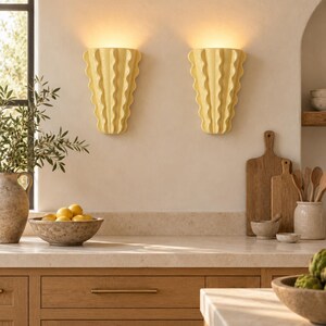 Puede incluir: Dos apliques de pared de cer&aacute;mica de color amarillo p&aacute;lido con un dise&ntilde;o vertical ondulado y plisado. Cada aplique est&aacute; montado en la pared y emite un brillo suave y c&aacute;lido desde la parte superior, proyectando luz hacia arriba.