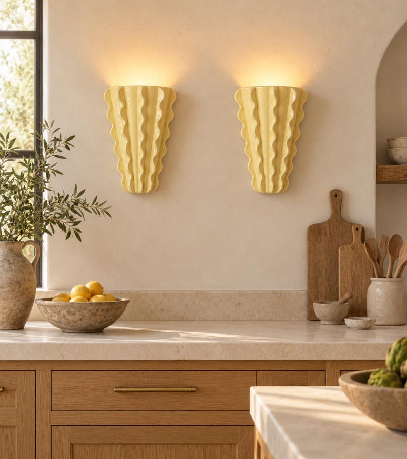Puede incluir: Dos apliques de pared de cer&aacute;mica de color amarillo p&aacute;lido con un dise&ntilde;o vertical ondulado y plisado. Cada aplique est&aacute; montado en la pared y emite un brillo suave y c&aacute;lido desde la parte superior, proyectando luz hacia arriba.
