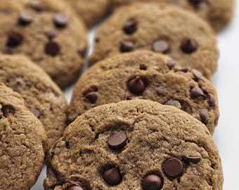 May include: Close-up of several freshly baked chocolate chip cookies. The cookies are a warm brown color, with visible chocolate chips scattered across their textured surfaces. The cookies are arranged on a white surface, suggesting a bakery or home-baked treat.