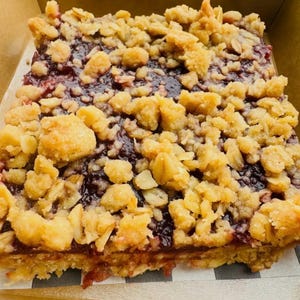 May include: A square fruit crumble bar featuring a thick layer of dark purple berry jam filling sandwiched between a golden brown oat-based crust and a generous topping of chunky, buttery oat streusel crumbles inside a cardboard box.