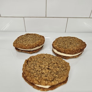 Puede incluir: Tres galletas de avena con relleno cremoso, dispuestas en un plato blanco. Las galletas son redondas y tienen una textura ligeramente rugosa.