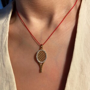 Op de afbeelding: Goudkleurige tennisracket-hangerketting met een rode koord. Het racket heeft een rasterpatroon en een rand van kleine, heldere stenen. De ketting wordt gedragen over een wit overhemd.