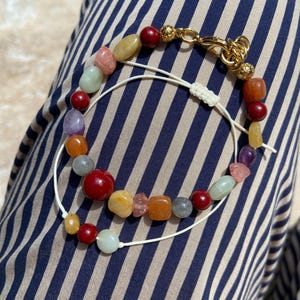 Puede incluir: Una pulsera de varias hebras con un surtido de cuentas de piedras preciosas pulidas e irregulares en tonos rojo, amarillo, naranja, morado y verde pálido. Las cuentas están ensartadas en un cordón blanco fino con un cierre de mosquetón dorado.