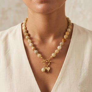 Könnte beinhalten: Eine Perlenkette aus runden, polierten Steinen in Erdtönen wie Beige, Hellbraun und Sandfarben. Die Kette verfügt über einen goldfarbenen Knebelverschluss in der Mitte, an dem eine kleine passende Steinperle und ein goldenes Herzanhänger hängen.