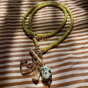 May include: A layered necklace with olive green beads and gold-colored accents. The necklace features a gold toggle clasp, a heart-shaped charm, a clear crystal, and a dalmatian jasper stone pendant.