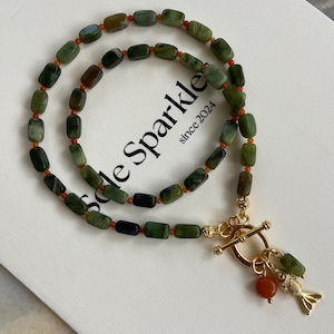 May include: A necklace featuring rectangular green stone beads interspersed with small orange beads. The piece includes a gold-toned toggle clasp with a dangling gold fish charm and a single round orange bead, arranged in a circular pattern.