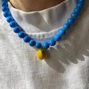May include: A blue beaded necklace with a yellow fruit pendant. The necklace features large, round, glossy blue beads. The pendant is a yellow fruit with a green leaf.
