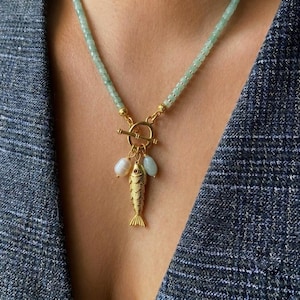 Può includere: Una collana con perline verde chiaro e una chiusura a T dorata. Il ciondolo è costituito da un ciondolo a forma di pesce dorato e testurizzato, affiancato da due piccole perle bianche irregolari, sospeso al centro della chiusura circolare.