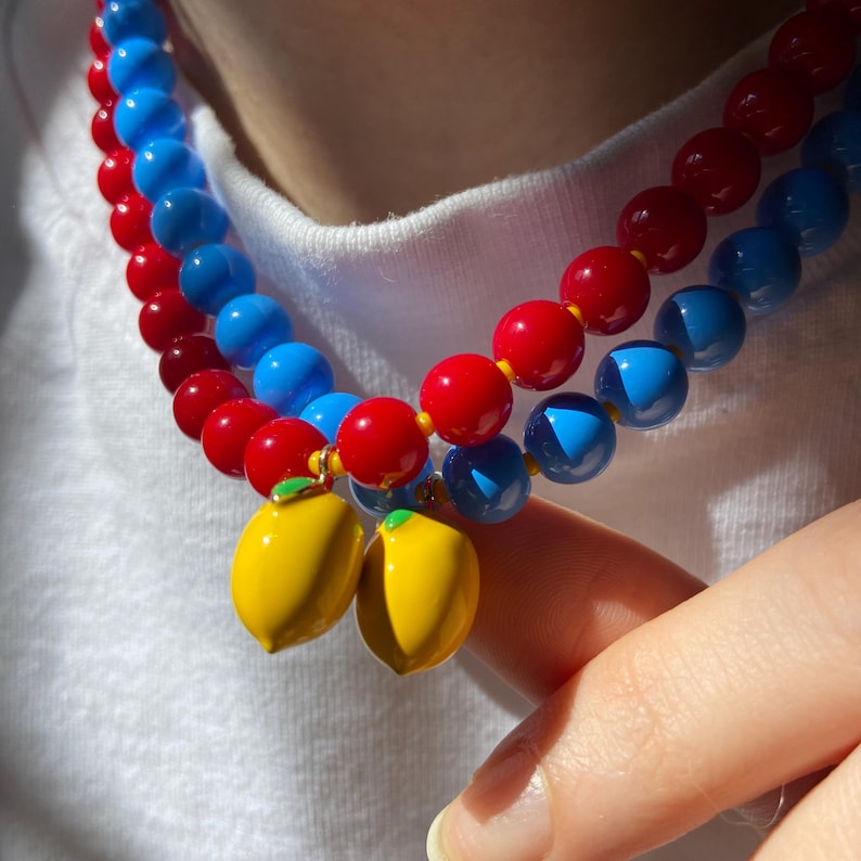 May include: A colorful necklace featuring red and blue beads, with a pair of yellow lemon charms. The necklace is composed of two strands of round beads, creating a vibrant and playful accessory.
