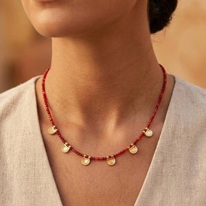 Puede incluir: Un collar que presenta una hilera de pequeñas cuentas rojas facetadas intercaladas con cinco dijes circulares de tono dorado espaciados uniformemente. Cada dije tiene un diseño semicircular texturizado en la parte superior y cuelga de la cadena.