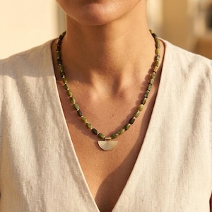 May include: A necklace featuring a strand of small, polished olive green stone beads interspersed with tiny gold-toned spacers. The necklace includes a central gold-toned pendant shaped like a semi-circle with a series of etched, concentric lines.