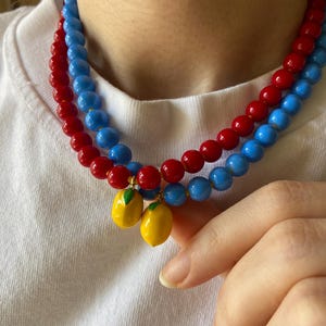 May include: A layered necklace featuring red and blue beaded strands. Two yellow lemon charms with green leaves dangle from the necklace. The necklace is worn against a white shirt.