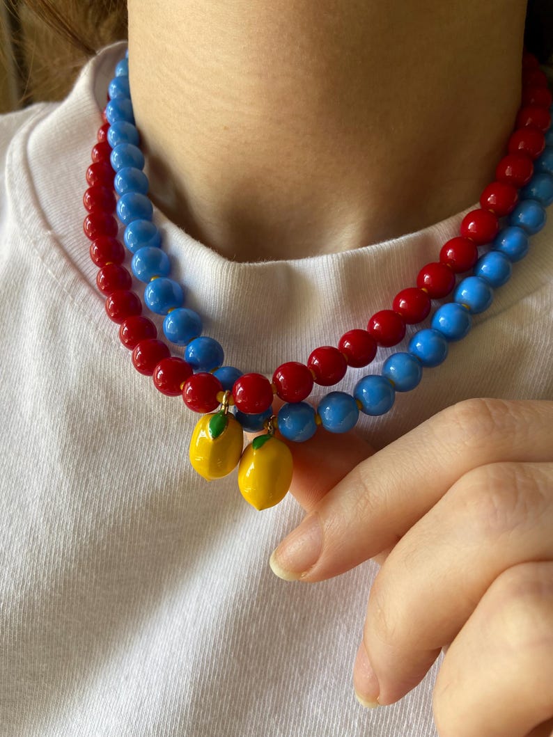 May include: A layered necklace featuring red and blue beaded strands. Two yellow lemon charms with green leaves dangle from the necklace. The necklace is worn against a white shirt.