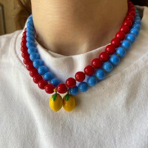 May include: Two-strand necklace featuring red and blue beads. The necklace has a yellow lemon charm with green leaves. The necklace is worn around the neck.