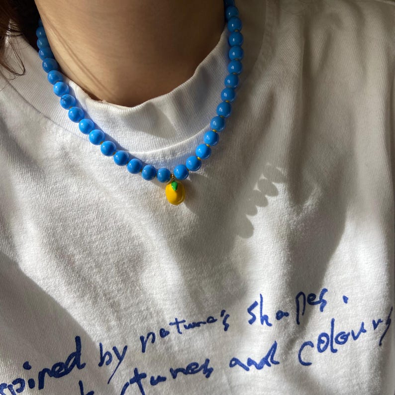 May include: A blue beaded necklace with a yellow and green lemon pendant. The necklace is worn over a white t-shirt with blue handwritten text that reads "inspired by nature's shapes, tunes and colour."