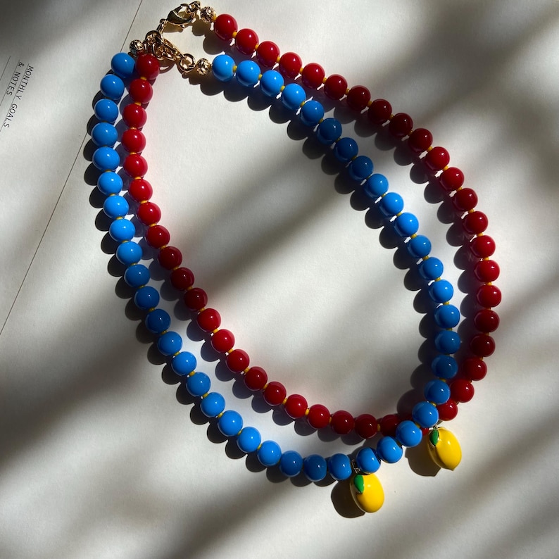 May include: Two beaded necklaces. One necklace is red and blue, the other is blue and red. Each necklace has a gold clasp and a yellow lemon charm with green leaves.