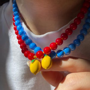 May include: Two beaded necklaces with red and blue beads. Each necklace has a yellow lemon charm with a green leaf. The necklaces are worn against a white shirt.