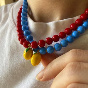 May include: Two beaded necklaces with red and blue beads. Each necklace has a lemon charm. The necklaces are worn over a white t-shirt.