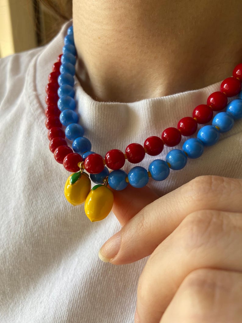 May include: Two beaded necklaces with red and blue beads. Each necklace has a lemon charm. The necklaces are worn over a white t-shirt.