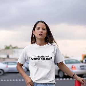 May include: A white t-shirt with the text "GENERATIONAL CURSE BREAKER" printed in black. The person wearing the shirt is standing in front of a red traffic cone.