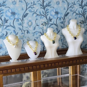 May include: Miniature jewelry display featuring four necklaces on white busts. Each necklace has a gold chain, with pendants including a pearl, a purple gem, and two dark gems. The display sits on a wooden shelf against a blue floral wallpaper.