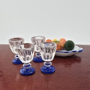 May include: Four clear glass goblets with blue bases on a wooden surface. The goblets are arranged in a row, with the bases facing the viewer. There is a white plate with fruit in the background.