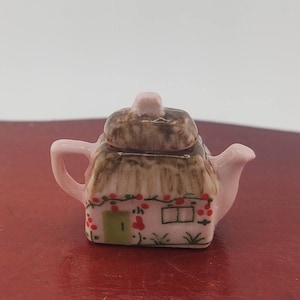 May include: A miniature pink teapot with a thatched roof design. The teapot features a small green door, a window, and red berry accents. The lid is brown, and the handle and spout are pink.