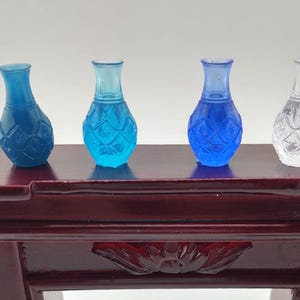 May include: Four decorative glass vases in different shades of blue and clear. The vases have a textured, diamond-like pattern and are arranged on a dark brown wooden surface.