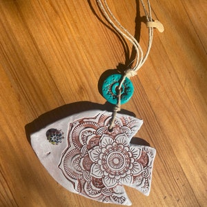 May include: A white ceramic fish ornament with a brown mandala design. The fish is hanging from a twine string with a turquoise ceramic bead.