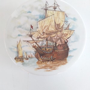 May include: A vintage ceramic plate with a hand-painted image of a large sailing ship with billowing sails, surrounded by smaller ships and a cloudy sky.