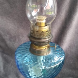 May include: A vintage oil lamp featuring a clear glass chimney and a blue, textured glass reservoir. The lamp has brass-coloured metal accents and a round base. The reservoir has a swirling pattern.