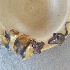 Peut inclure: Un plat en céramique avec une finition beige clair mouchetée. Le plat est décoré d'un groupe de feuilles marron et vertes et d'une petite figurine de souris brune. La souris a une longue queue incurvée.
