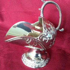 May include: A silver sugar bowl with a handle and floral embellishments. The bowl has a scoop shape and rests on a decorative base. The handle curves upwards, enhancing the elegant design. The bowl is likely made of silver-plated metal.