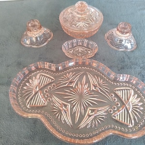 Könnte beinhalten: Eine Sammlung von rosa Glasserviceteilen, darunter ein Tablett, eine Schale mit Deckel, zwei Kerzenhalter und eine kleine Schale. Die Teile haben ein detailliertes, geätztes Design. Das Tablett ist länglich mit einem gewellten Rand.