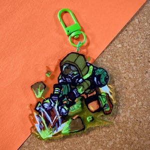 May include: A colorful acrylic charm featuring a cartoon character in a gas mask and protective gear. The charm has a green clasp and is set against an orange and brown background. The character is primarily green, with accents of orange, white, and black.