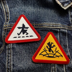 May include: Two embroidered patches with red borders are displayed on a denim jacket. One patch is a white triangle with a black figure crossing a dotted line. The other is a yellow triangle with a falling figure.