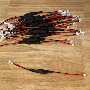 Op de afbeelding: Een verzameling zwarte en rode elektrische draden met witte connectoren. De draden zijn gebundeld en verspreid over een houten oppervlak. De draden hebben zwarte krimpkous in het midden.