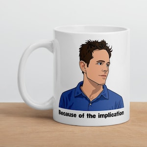 May include: White ceramic mug with a cartoon illustration of a man in a blue shirt. The mug has the text "Because of the implication" printed below the illustration. The mug is on a wooden surface.