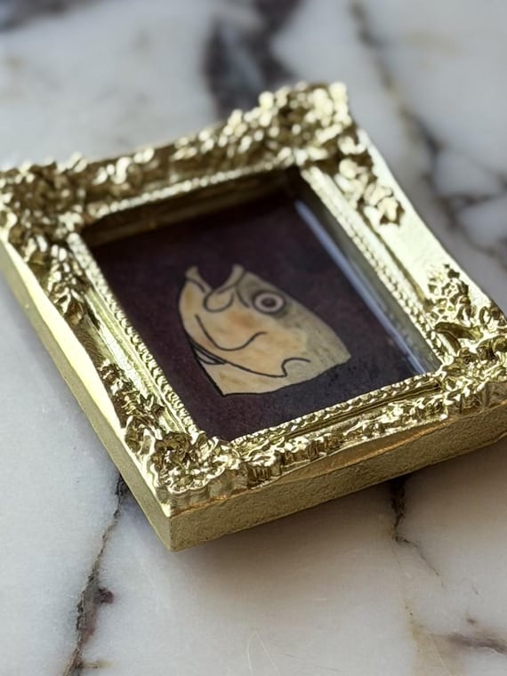 A tiny ornate-framed fish head portrait that treats seafood with the gravitas of a Renaissance painting — absurdist miniature art for your fridge.
