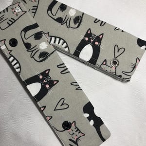 May include: Two gray fabric wristlets with black and white cat designs and pink hearts. The wristlets have black buttons and the text "Pr 3:6" is printed on one of the wristlets.