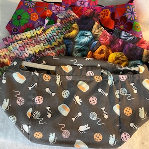 Può includere: Una collezione di borse e pochette in tessuto con vari motivi. Le borse presentano disegni floreali, di filato e a tema gatto. I colori includono viola, arancione, rosa e grigio. Le borse sono probabilmente per trasportare forniture per maglieria o altri piccoli oggetti.