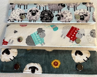May include: Three rectangular fabric strips with metal snap buttons. The top strip is light blue with white and black sheep. The middle strip is white with sheep wearing red and blue sweaters. The bottom strip is dark green with white sheep and sunflowers.