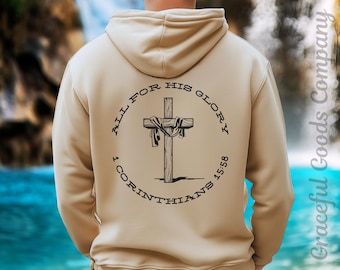 May include: Beige hoodie with a black graphic on the back featuring a cross, draped fabric, and the text "ALL FOR HIS GLORY" and "1 CORINTHIANS 15:58". The hoodie is a neutral color and has a drawstring hood.
