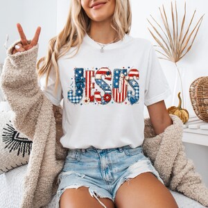 Könnte beinhalten: Weißes T-Shirt mit dem Wort "JESUS" in einem patriotischen Design mit roten, weißen und blauen Mustern. Die Buchstaben enthalten Sterne, Streifen und Vichy-Karo und schaffen ein festliches und religiöses Thema. Das Shirt wird mit Jeansshorts kombiniert.